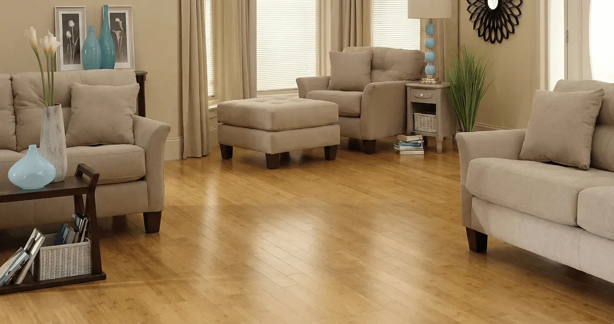 A living room with professionally installed bamboo flooring, beige sofas, a matching ottoman, a side table with a blue lamp, vases, framed pictures, and large windows with blinds and curtains in Peoria, IL